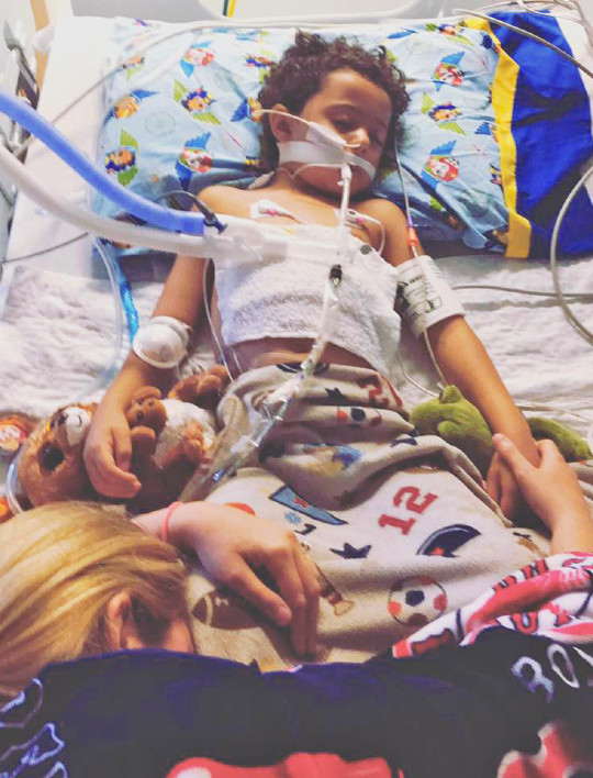a boy lying in a hospital bed with lots of tubes and his mother draped over him