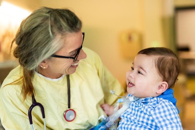 A woman wearing a stethoscope and a young patient on a ventilator look lovingly at one another.